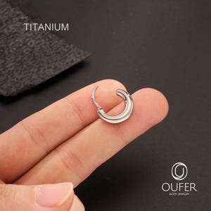 Puede incluir: Un solo pendiente de aro de titanio plateado brillante. El pendiente est&aacute; abierto, revelando el cierre. La palabra "Titanium" est&aacute; impresa en blanco en la esquina superior izquierda. El pendiente est&aacute; sujeto por un dedo.