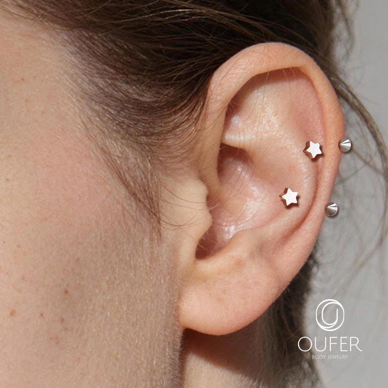 May include: Close-up of an ear with two star-shaped earrings and two silver-colored spike earrings. The earrings are small and delicate, adding a touch of sparkle. The ear is smooth and the skin is light-toned. The image is well-lit, highlighting the jewelry.