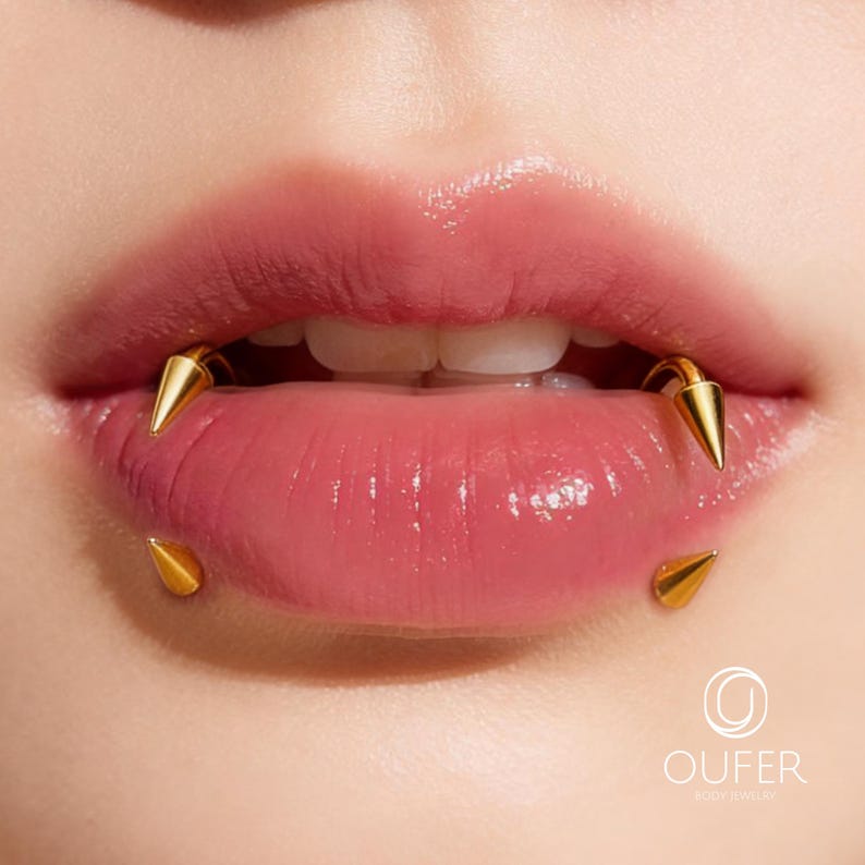 May include: A set of gold-toned spike lip piercings featuring two curved barbells with pointed ends positioned on the upper and lower lips. The jewelry has a polished metallic finish. The logo OUFER BODY JEWELRY is visible in the bottom corner.