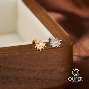 Könnte beinhalten: Zwei Labret-Piercings mit flacher Rückseite und einem Sonnenstrahlen-Design mit einem zentralen, birnenförmigen, klaren Edelstein. Ein Stecker ist goldfarben, der andere silberfarben. Jedes Stück hat ein strahlenförmiges Muster um den Stein.
