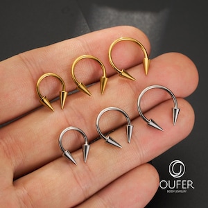 May include: Six circular horseshoe-shaped body jewelry pieces with pointed spike ends. Three are gold-toned and three are silver-toned. The jewelry is arranged in two rows. The text OUFER BODY JEWELRY and a circular logo are visible in the corner.