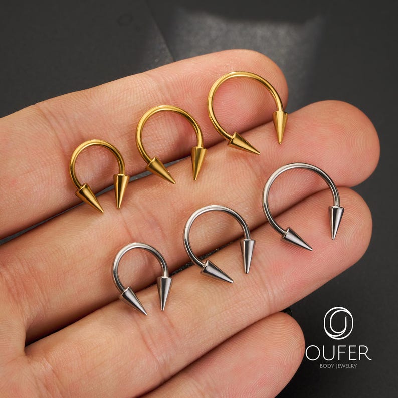 May include: Six circular horseshoe-shaped body jewelry pieces with pointed spike ends. Three are gold-toned and three are silver-toned. The jewelry is arranged in two rows. The text OUFER BODY JEWELRY and a circular logo are visible in the corner.