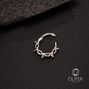 Op de afbeelding: Een zilverkleurige scharnierende oorring met een kettingachtig ontwerp, geaccentueerd door gelijkmatig verdeelde, scherpe, conische spikes. De oorring is open, waardoor het scharniermechanisme zichtbaar is. De achtergrond is donker en getextureerd.