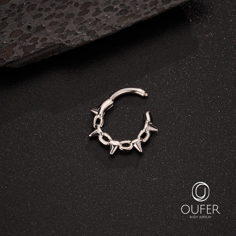 Op de afbeelding: Een zilverkleurige scharnierende oorring met een kettingachtig ontwerp, geaccentueerd door gelijkmatig verdeelde, scherpe, conische spikes. De oorring is open, waardoor het scharniermechanisme zichtbaar is. De achtergrond is donker en getextureerd.