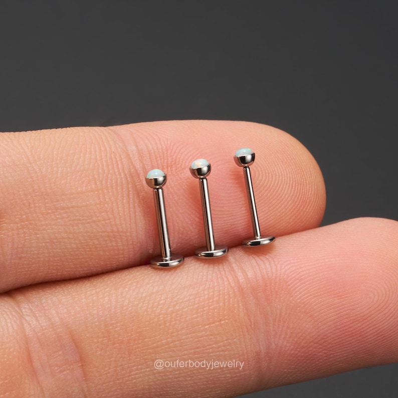 Op de afbeelding: Drie zilveren barbell oorbellen met opaalachtige witte stenen. De oorbellen worden op een vinger getoond.