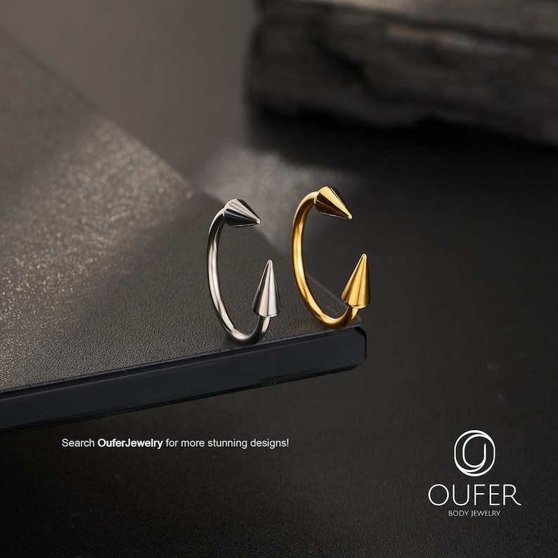 May include: Two circular body jewelry rings, one silver-toned and one gold-toned, each featuring two pointed cone-shaped spikes at the ends. The text "Search OuferJewelry for more stunning designs!" and the "OUFER BODY JEWELRY" logo are visible.
