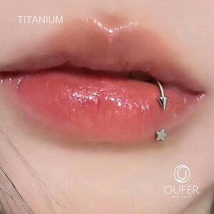 May include: Close-up of a person's lips with a silver lip ring and a star-shaped stud piercing. The word "TITANIUM" is visible in the upper left corner. The lips are glossy and a natural pink color.