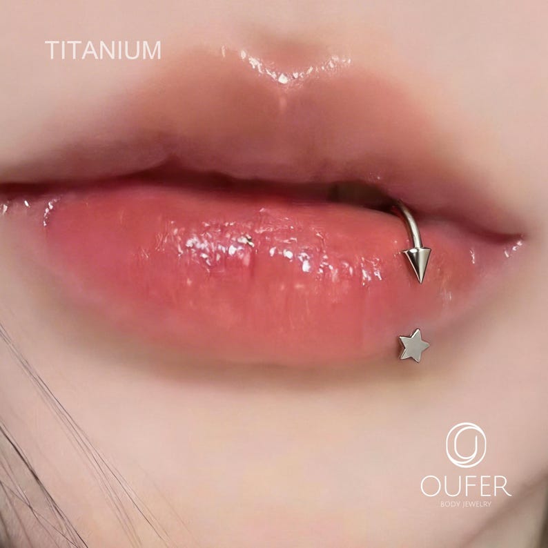 May include: Close-up of a person's lips with a silver lip ring and a star-shaped stud piercing. The word "TITANIUM" is visible in the upper left corner. The lips are glossy and a natural pink color.