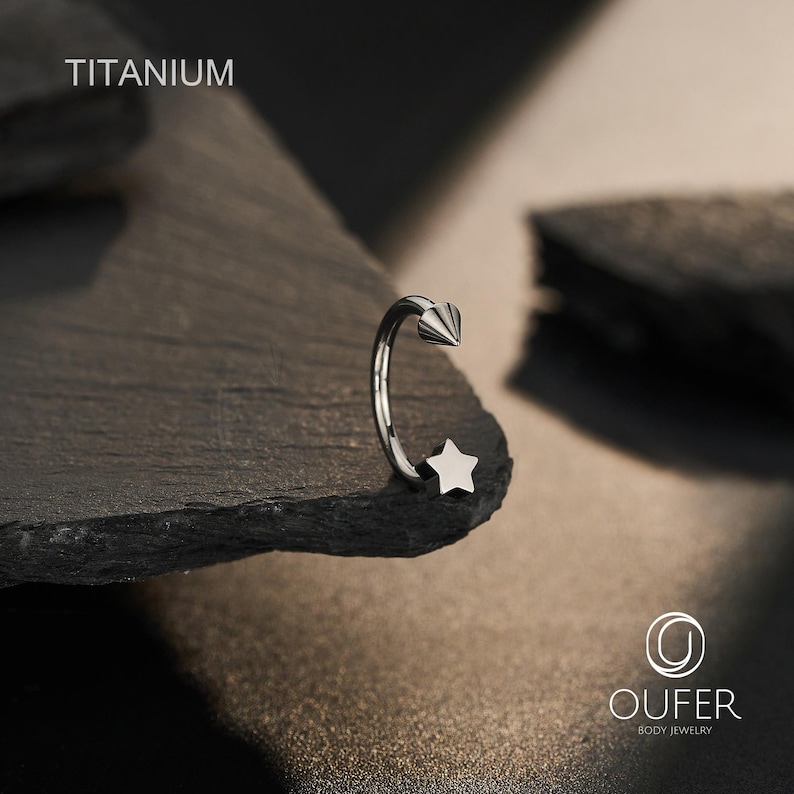May include: A silver-colored body jewelry ring with a star and cone-shaped design. The word "TITANIUM" is printed at the top. The ring is displayed on a dark, textured surface. The brand logo "OUFER BODY JEWELRY" is at the bottom.