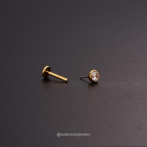 May include: A gold body jewelry piercing with a clear round gem. The piercing is shown with the post removed.