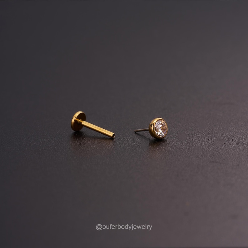 May include: A gold body jewelry piercing with a clear round gem. The piercing is shown with the post removed.