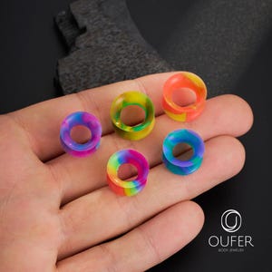 May include: Five colorful silicone ear gauges in various rainbow hues, including purple, green, orange, and blue. The gauges are circular with a hollow center, and are displayed on a hand. The logo for Oufer Body Jewelry is in the bottom right corner.