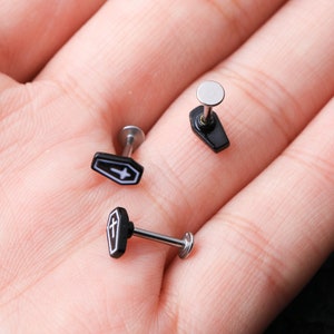 May include: Three silver and black coffin-shaped body jewelry pieces. Two of the pieces have a small cross design on the coffin. The third piece is a plain silver stud.
