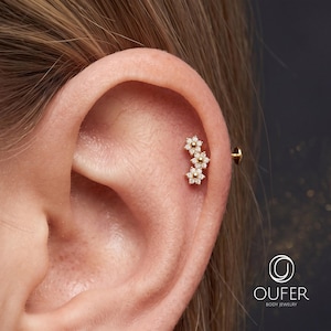Può includere: Un orecchino da cartilagine color oro con tre ciondoli collegati a forma di fiore, ciascuno incastonato con piccole pietre trasparenti. L'orecchino è fissato con un perno a retro piatto. Il logo OUFER BODY JEWELRY è visibile in basso a destra.