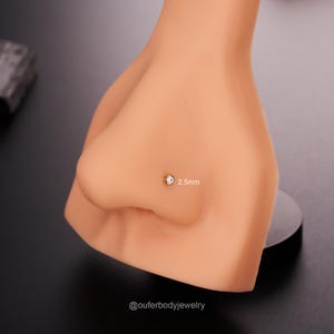 May include: A close-up of a clear nose stud with a small, round, clear gemstone. The stud is 2.5mm in size and is shown on a model's nose.