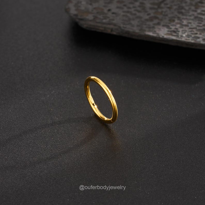 May include: A gold-colored seamless ring, a type of body jewelry, is shown on a dark gray surface. The ring is made of surgical steel and is designed for piercing.