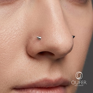 May include: A silver-colored nose piercing featuring a sharp, conical spike stud. The jewelry is positioned on the side of the nostril. The image includes the text OUFER BODY JEWELRY with a circular logo icon in the bottom right corner.