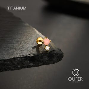 May include: A gold-tone titanium flat back stud earring featuring a round pink stone and a smaller clear cubic zirconia stone. The text TITANIUM is visible in the upper left corner and the OUFER BODY JEWELRY logo is in the bottom right corner.
