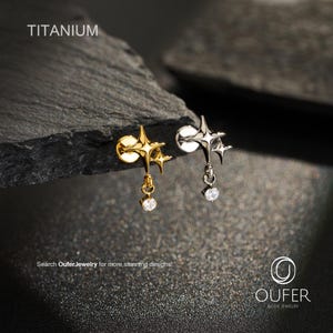 Pode incluir: Dois brincos de titânio com um design de estrela de quatro pontas e uma pedra preciosa redonda transparente pendurada. Um brinco é dourado e o outro prateado. O texto TITANIUM e OUFER BODY JEWELRY é visível na imagem.