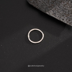 Puede incluir: Un anillo de piercing corporal sin costuras de plata. El anillo es liso y brillante, y se muestra sobre un fondo negro.