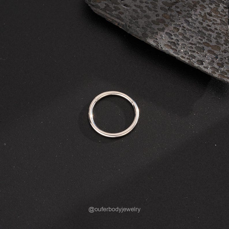 Puede incluir: Un anillo de piercing corporal sin costuras de plata. El anillo es liso y brillante, y se muestra sobre un fondo negro.