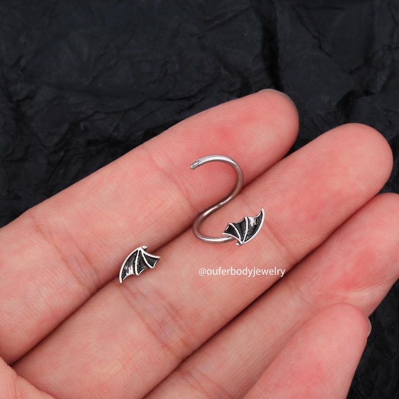 Peut inclure: Deux boucles d'oreilles en h&eacute;lice argent&eacute;es avec des motifs d'ailes de chauve-souris noires. Les boucles d'oreilles sont pr&eacute;sent&eacute;es sur une main, la plus grande sur l'index et la plus petite sur le pouce. Le texte '@ouferbodyjewelry' est visible sur l'image.