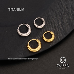 Puede incluir: Cuatro pendientes de aro de titanio en tonos plateados y dorados se muestran sobre una superficie oscura. La palabra "TITANIUM" est&aacute; impresa en la parte superior. El texto "Search OuferJewelry for more stunning designs!" est&aacute; en la parte inferior, junto con el logotipo de Oufer Body Jewelry.