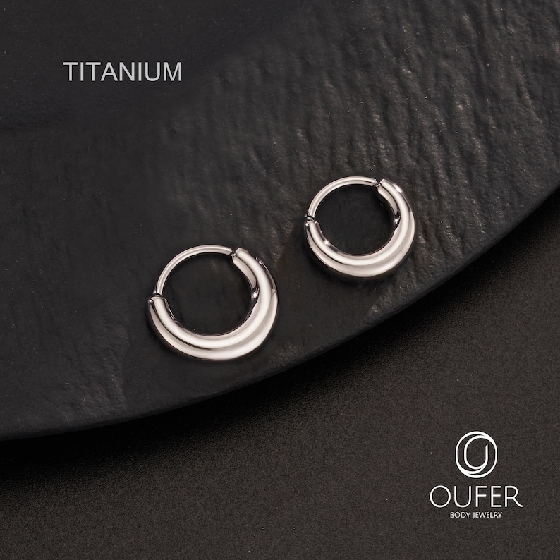Puede incluir: Dos pendientes de aro de titanio plateado. Los pendientes tienen forma de media luna y un acabado pulido. La palabra "TITANIUM" est&aacute; impresa en blanco en la parte superior izquierda. Los pendientes se muestran sobre una superficie oscura. La marca "OUFER BODY JEWELRY" est&aacute; impresa en la parte inferior derecha.