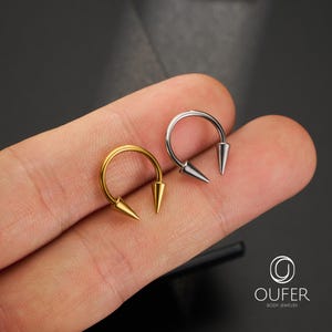 May include: Two circular horseshoe-shaped body jewelry pieces, one in a gold tone and one in a silver tone, each featuring two pointed cone-shaped ends. The jewelry is displayed against a dark background with the text OUFER BODY JEWELRY.