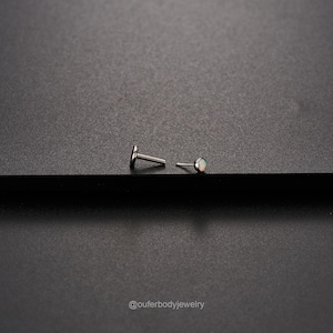 Op de afbeelding: Een paar zilveren body jewelry studs met een kleine opaal edelsteen op een stud. De studs liggen op een zwart oppervlak.