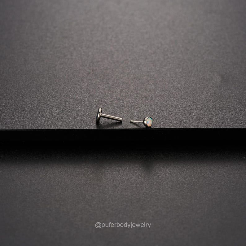 Op de afbeelding: Een paar zilveren body jewelry studs met een kleine opaal edelsteen op een stud. De studs liggen op een zwart oppervlak.