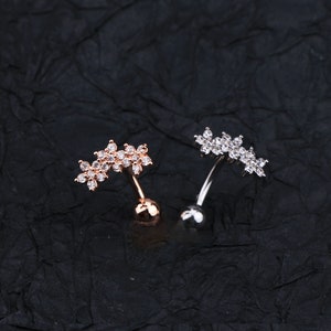 May include: Two belly button rings with a flower design. The ring on the left is rose gold with clear stones. The ring on the right is silver with clear stones.