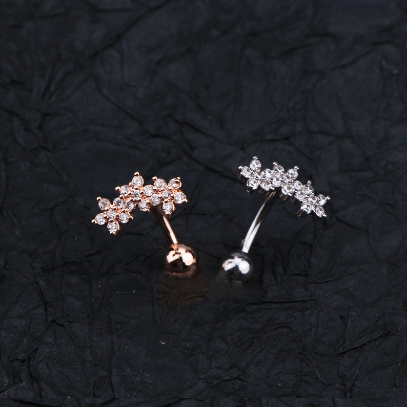 May include: Two belly button rings with a flower design. The ring on the left is rose gold with clear stones. The ring on the right is silver with clear stones.
