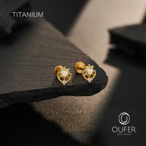 Könnte beinhalten: Ein Paar goldfarbene Titan-Ohrstecker mit einem sonnenstrahlenförmigen Design, einem zentralen runden weißen Opal-Cabochon und einem kleinen klaren Kristall an der Basis. Der Text TITANIUM und OUFER BODY JEWELRY ist im Bild sichtbar.