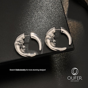 May include: A pair of silver-toned hoop earrings featuring a crescent moon design with a detailed human face embossed on the surface. The earrings include a post-back closure and are branded with the text Oufer Jewelry in the corner.