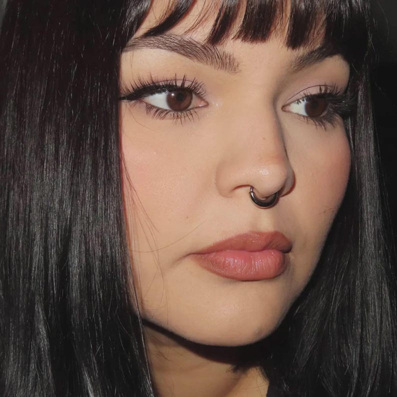 Puede incluir: Primer plano del rostro de una persona con cabello oscuro y un piercing en el tabique nasal. La persona tiene ojos marrones, pesta&ntilde;as largas y usa labial. El fondo es oscuro.