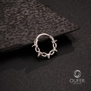 Op de afbeelding: Een zilverkleurige lichaamsjuwelenring met een ketting- en spijkerontwerp. De ring is cirkelvormig met een scharniersluiting. De schakels van de ketting zijn verbonden met kleine spikes. De sieraden worden getoond op een donker, getextureerd oppervlak.