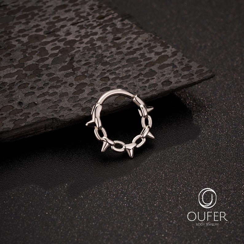 Op de afbeelding: Een zilverkleurige lichaamsjuwelenring met een ketting- en spijkerontwerp. De ring is cirkelvormig met een scharniersluiting. De schakels van de ketting zijn verbonden met kleine spikes. De sieraden worden getoond op een donker, getextureerd oppervlak.