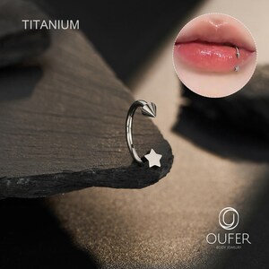 May include: A silver-colored titanium lip ring with a star and cone design. The word "TITANIUM" is printed at the top. A close-up of a lip with a similar ring is in the upper right corner. The brand name "OUFER BODY JEWELRY" is at the bottom.