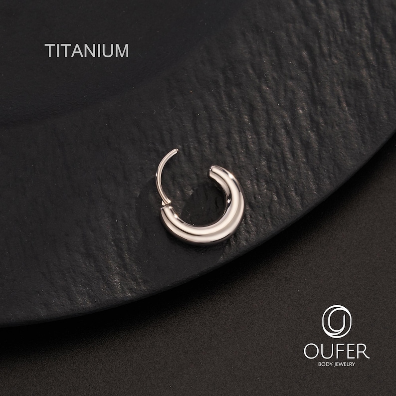 Puede incluir: Pendiente de aro de titanio plateado. El pendiente tiene un dise&ntilde;o peque&ntilde;o y curvado con un cierre de bisagra. La palabra "TITANIUM" est&aacute; impresa en blanco en la parte superior izquierda. Se muestra sobre una superficie negra texturizada.