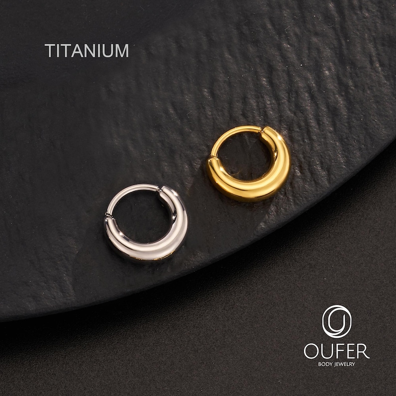 Puede incluir: Dos pendientes de aro de titanio, uno plateado y otro dorado, se muestran sobre una superficie oscura. La palabra "TITANIUM" est&aacute; impresa encima de los pendientes. Los pendientes tienen un dise&ntilde;o simple y redondeado.