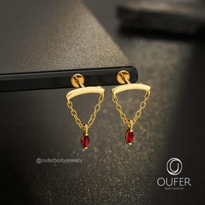 Puede incluir: Dos pendientes dorados de parte posterior plana con una barra curva conectada a una cadena delicada que suspende una pequeña piedra preciosa roja de corte marquesa. La imagen incluye el texto @ouferbodyjewelry y el logotipo OUFER BODY JEWELRY.