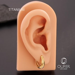 Puede incluir: Un pendiente de aro dorado se muestra en un modelo de oreja color carne. La palabra "TITANIUM" es visible. El pendiente tiene un dise&ntilde;o liso y curvado. El logotipo de la marca "OUFER BODY JEWELRY" est&aacute; en la esquina inferior derecha. De titanio.