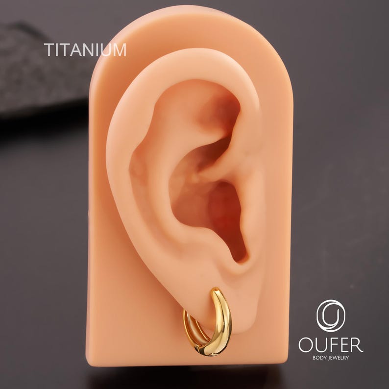 Puede incluir: Un pendiente de aro dorado se muestra en un modelo de oreja color carne. La palabra "TITANIUM" es visible. El pendiente tiene un dise&ntilde;o liso y curvado. El logotipo de la marca "OUFER BODY JEWELRY" est&aacute; en la esquina inferior derecha. De titanio.