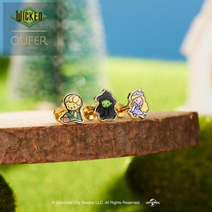 May include: Three gold-tone stud earrings featuring colourful enamel character designs. One earring depicts a blonde figure in green, one shows a green-skinned figure in a black dress, and one displays a blonde figure in a light purple dress.