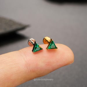 Può includere: Un paio di piccoli orecchini a forma triangolare con pietre preziose verdi. Uno ha un perno d'argento, l'altro d'oro. Gli orecchini sono mostrati su un dito contro uno sfondo scuro. Il testo "@ouferbodyjewelry" è visibile.
