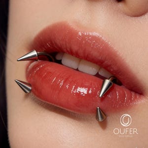 May include: A silver-toned metal lip piercing jewelry piece featuring three sharp, conical spikes. The jewelry is positioned around the lips, with the curved bar passing through the lip tissue. The OUFER BODY JEWELRY logo is visible in the corner.