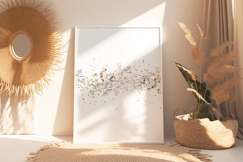 May include: A white framed print with a watercolor illustration of a branch of white flowers with gold glitter accents. The print is hanging on a white wall with a woven sunburst mirror and a wicker basket with dried plants.