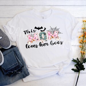 May include: White t-shirt with the text "This MOM loves her boos" in a playful font, decorated with bats, spiderwebs, and ghosts. The shirt is paired with blue jeans and gray sneakers, suggesting a casual, everyday style.
