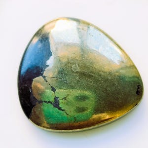 May include: A polished, teardrop-shaped stone with a metallic sheen. The stone features a blend of colors, including gold, green, blue, and black, with a cracked texture across its surface. The stone is set against a white background.
